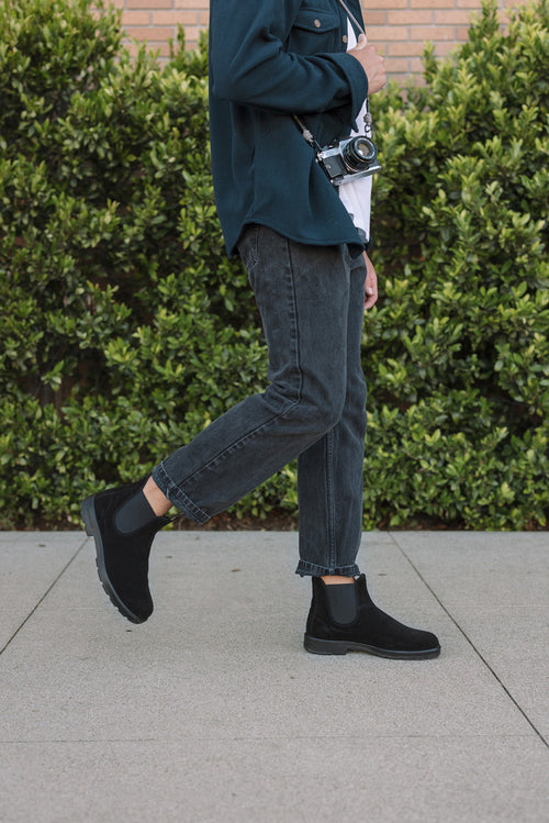 A person wearing black boots, cropped dark jeans, and a dark jacket walks on a sidewalk holding a camera, with green shrubs and a brick wall in the background.
