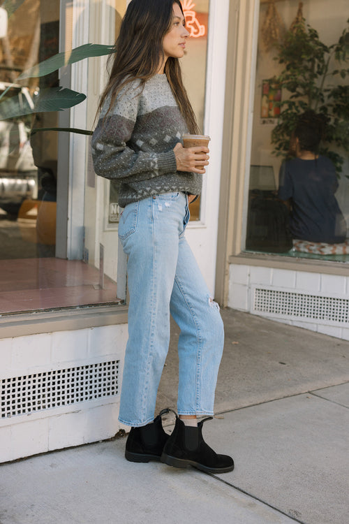 A woman with long hair stands outside a cafe holding a coffee cup, wearing a gray patterned sweater, light blue ripped jeans, and black boots. Another person sits inside the cafe by the window.
