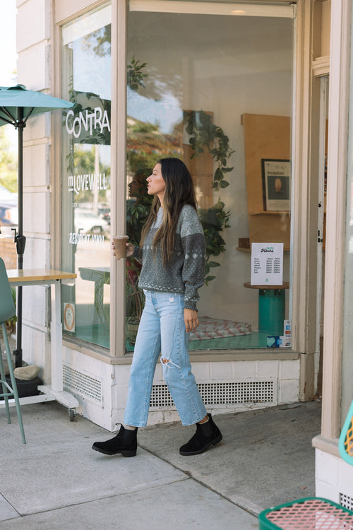 A woman with long dark hair, wearing a gray sweater, ripped light blue jeans, and black boots, holds a drink while walking out of a cafe with glass windows and green outdoor chairs.