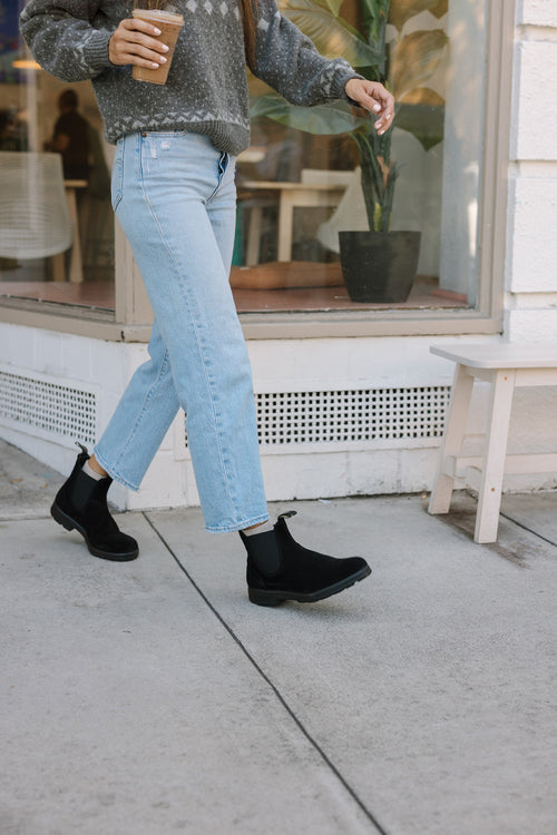 A person wearing light blue jeans, black boots, and a gray sweater walks on a sidewalk while holding a cup of coffee, passing by a window with a potted plant inside.