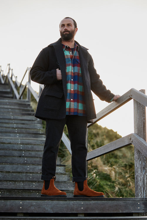 A bearded man in a black coat, colorful plaid shirt, dark jeans, and brown boots stands on outdoor wooden stairs, holding the railing and looking into the distance.