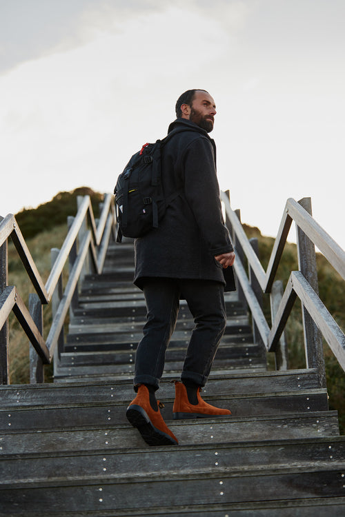 A man wearing a dark coat, jeans, and brown boots with a black backpack walks up wooden stairs outdoors, looking back over his shoulder. The sky is cloudy and grassy hills are in the background.