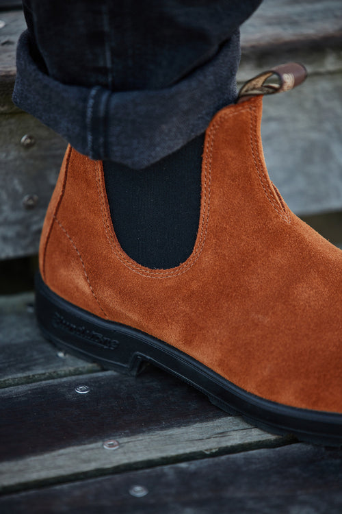 A close-up of a person wearing dark jeans and a rust-colored suede Chelsea boot with a black elastic side panel, standing on wooden planks.