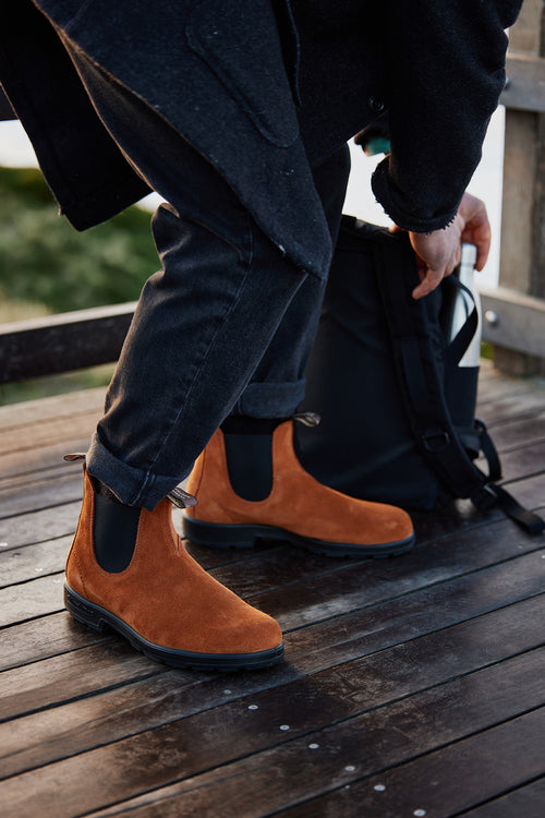 A person in black jeans and a dark coat adjusts their brown suede Chelsea boots while crouching beside a black backpack on a wooden deck.
