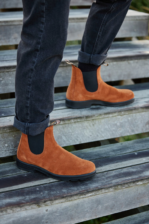 A person wearing rolled-up black jeans and tan suede Chelsea boots is walking up weathered wooden steps.