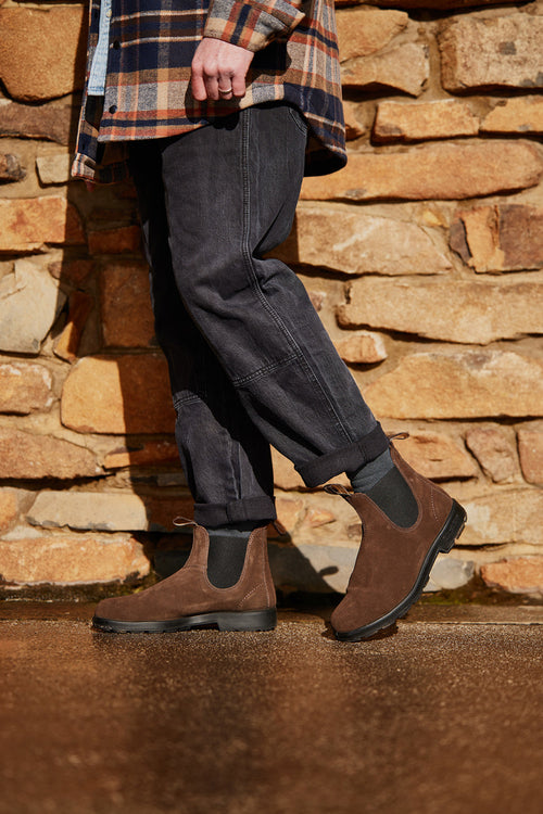 A person wearing dark jeans, a plaid shirt, and brown suede Chelsea boots stands on a wet surface in front of a stone wall. Only the lower half of the body is visible.
