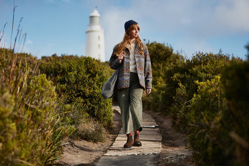 A woman wearing a patterned jacket, green pants, and a beanie walks along a narrow path surrounded by bushes, with a white lighthouse visible in the background under a partly cloudy sky.