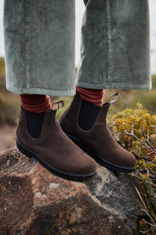 A person wearing brown Chelsea boots, red socks, and green corduroy pants stands on a large rock outdoors, surrounded by greenery.