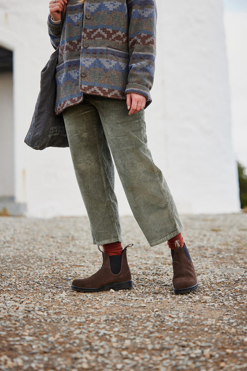 A person stands outdoors on gravel, wearing olive green corduroy pants, a patterned jacket, brown Chelsea boots, red socks, and holding a dark tote bag. The background is white and out of focus.