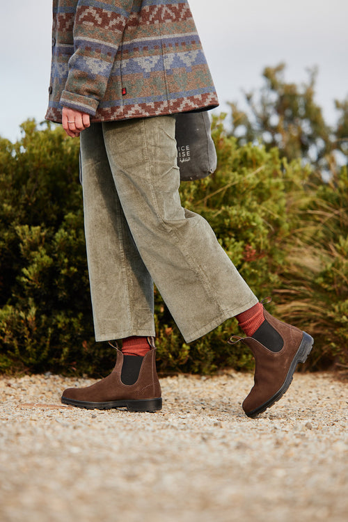 A person wearing olive green pants, a patterned jacket, red socks, and brown Chelsea boots walks on a gravel path with green bushes in the background.