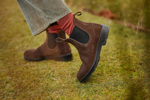 Person wearing brown suede Chelsea boots with black elastic panels, red socks, and light brown corduroy pants, standing on green grass outdoors.