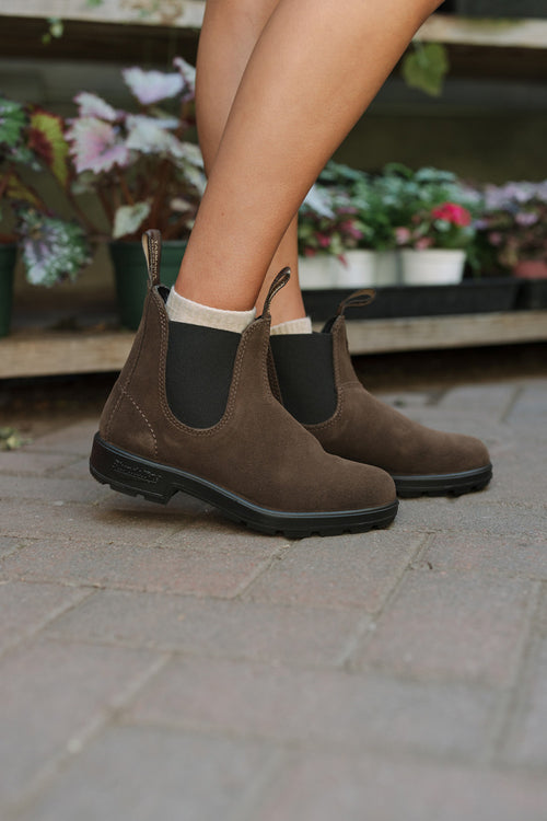 A person wearing brown suede Chelsea boots with black elastic sides and cream socks stands on a paved surface near potted plants.