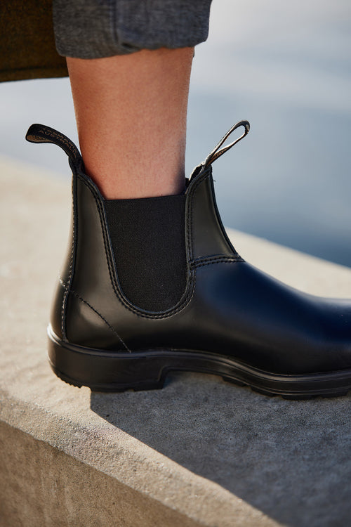 A close-up of a person wearing a black leather Chelsea boot, standing on the edge of a concrete surface with water blurred in the background. The person’s pants are cuffed at the ankle.