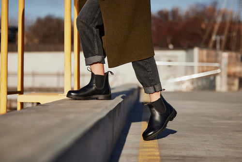 A person wearing cuffed black jeans, a long olive coat, and black Chelsea boots steps up onto a concrete curb outdoors on a sunny day.