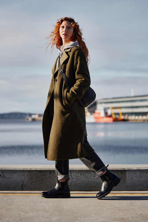 A person with curly red hair wearing a long olive green coat, black boots, cuffed jeans, and a gray crossbody bag walks along a waterfront with modern buildings and water in the background.