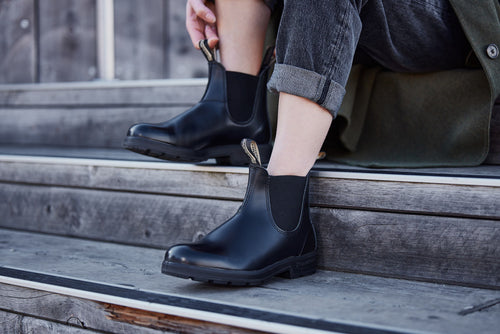 A person sits on wooden steps, wearing rolled-up dark jeans and shiny black Chelsea boots, preparing to stand up or adjust their boots.