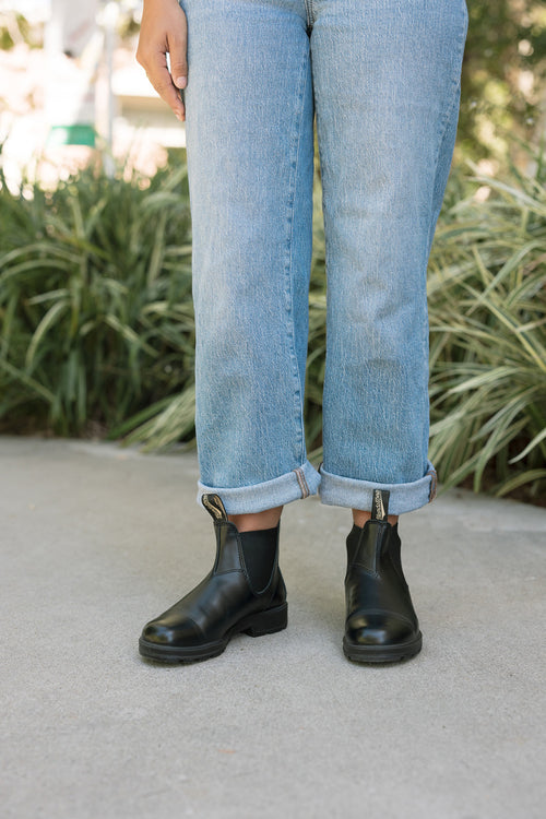 A person stands on a sidewalk with rolled-up blue jeans and black ankle boots. Green plants are in the background.