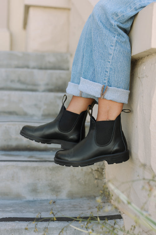 A person wearing cuffed light blue jeans and black Chelsea boots sits on outdoor concrete steps, with their legs extended and feet resting on a higher step.