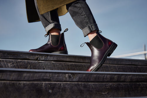 A person wearing dark red Chelsea boots and cuffed black jeans walks up wooden stairs outdoors on a sunny day.
