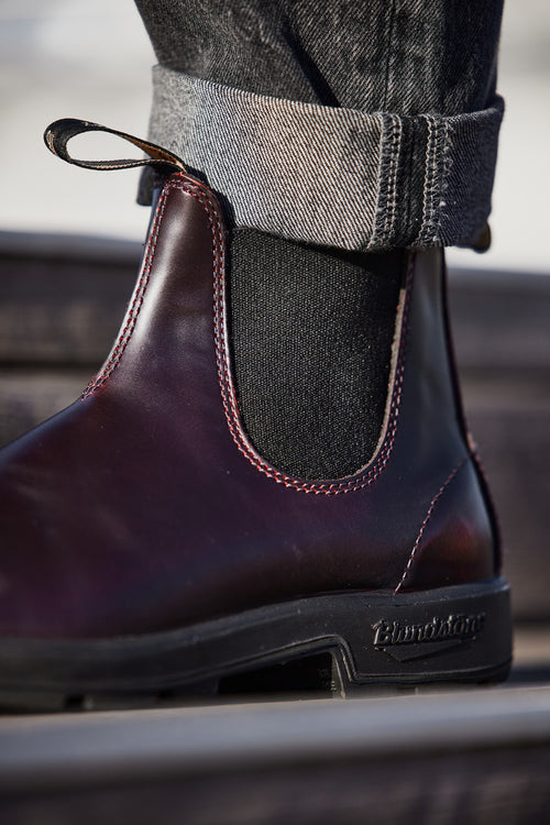 A close-up of a person wearing a dark burgundy Blundstone Chelsea boot, black socks, and rolled-up dark jeans. The focus is on the side of the boot, highlighting its stitching and logo on the sole.