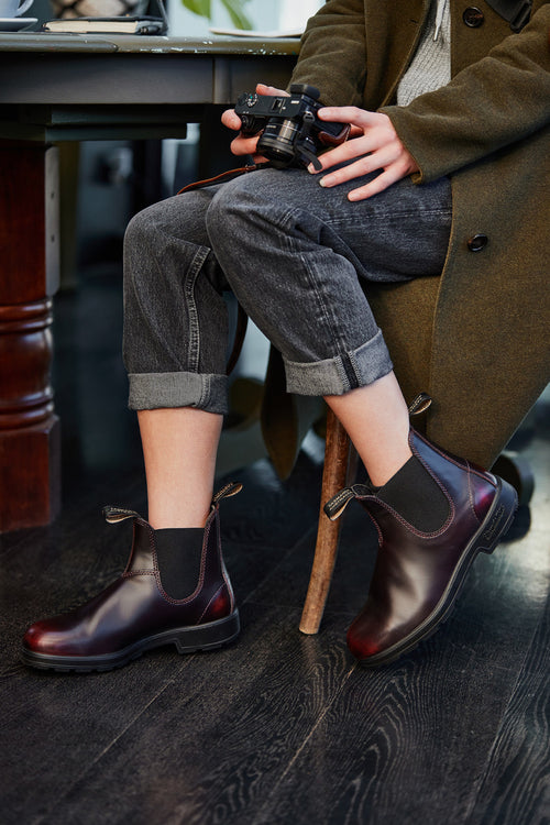 A person sitting on a chair, holding a camera, wearing rolled-up dark jeans, an olive green coat, and dark brown Chelsea boots with black elastic sides. Only the lower half of their body is visible.