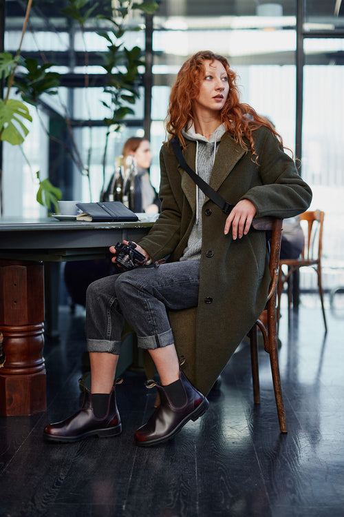 A person with long, wavy red hair sits on a chair in a modern indoor space, wearing a green coat, grey pants, and dark boots, holding a camera and looking to the side. Books and plants are on the table nearby.