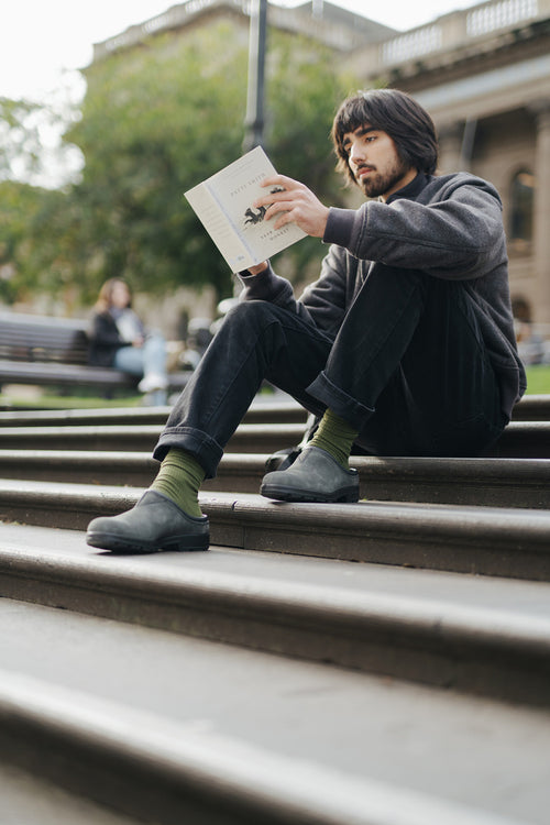 A man with dark hair and a beard sits on outdoor steps, reading a book. He wears a dark sweater, black pants, green socks, and black shoes. Blurred buildings and greenery appear in the background.