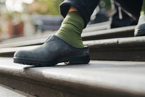 A person wearing green socks and black slip-on shoes is stepping on a set of outdoor stairs. Only their lower leg and foot are visible.