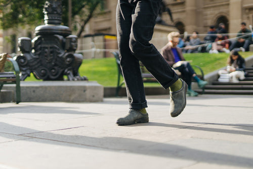 Person wearing dark jeans and green socks with gray shoes walking briskly on a paved sidewalk in an urban park, with blurred people sitting on steps and greenery in the background.