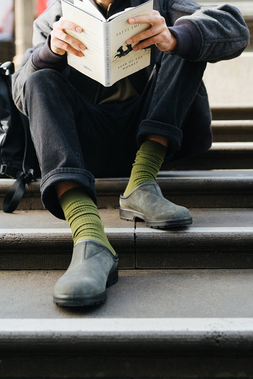 A person sitting on outdoor steps, wearing green socks and black slip-on shoes, is reading a book titled M Train by Patti Smith. Their face is not visible in the image.