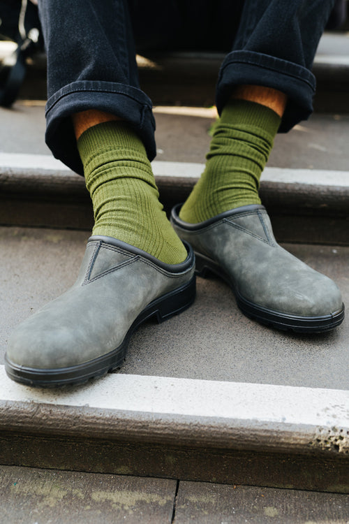 A person wearing green ribbed socks and gray slip-on clogs sits on outdoor steps, with black pants slightly cuffed above the ankles.