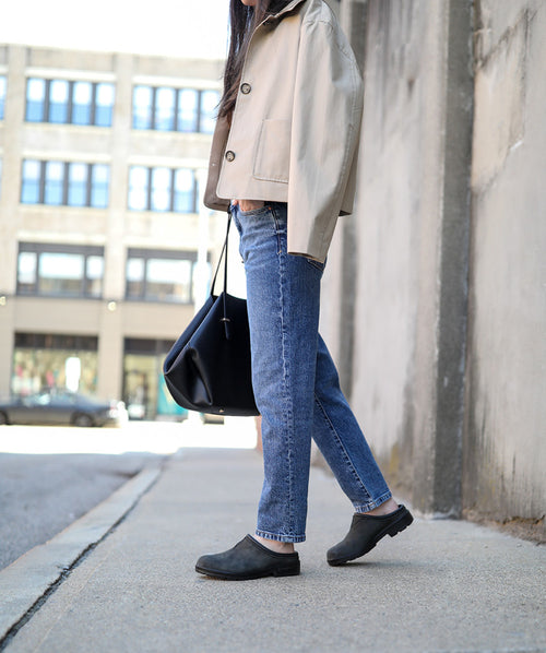 A person stands on a sidewalk wearing blue jeans, a beige jacket, black slip-on shoes, and carrying a black bag. Their head is not visible, and an urban building is in the background.