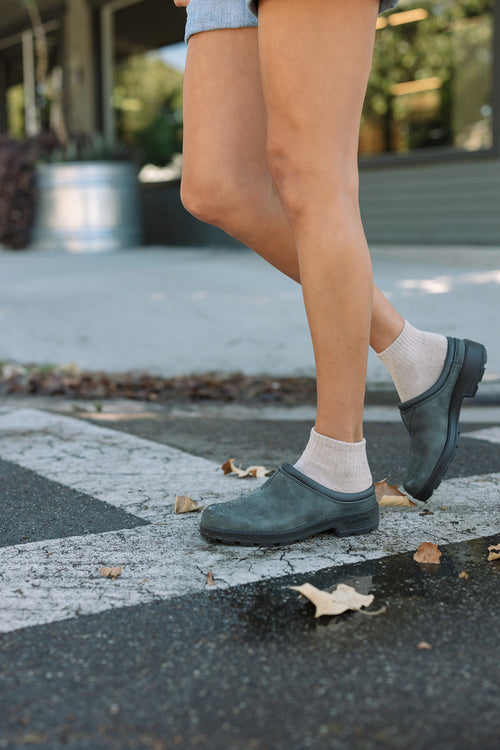 A person wearing light socks and black clogs stands on a crosswalk, with dry leaves scattered on the ground. Only their lower legs are visible.