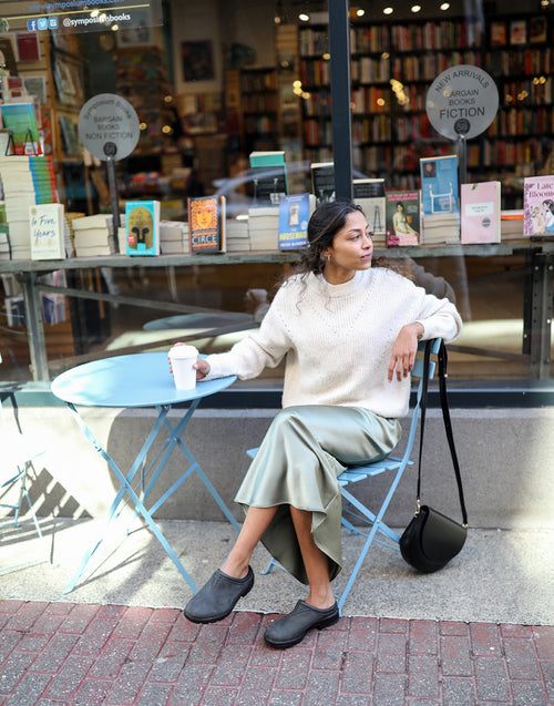 A woman sits at a blue outdoor table in front of a bookstore, holding a coffee cup. She wears a white sweater, light green skirt, and black shoes, with a black purse hanging on the chair. Books are displayed in the shop window behind her.