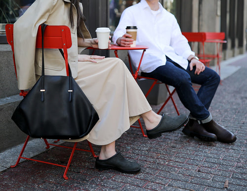 Two people sit at a red outdoor café table, holding coffee cups. One wears a tan coat and dress with black clogs, and has a large black handbag. The other wears a white shirt, dark jeans, and black shoes.
