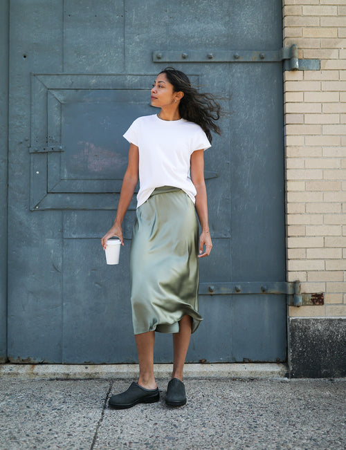 A woman stands outdoors against a large blue metal door, wearing a white t-shirt, light green satin skirt, and black shoes. She holds a white cup and looks to the side, with her hair blowing in the wind.