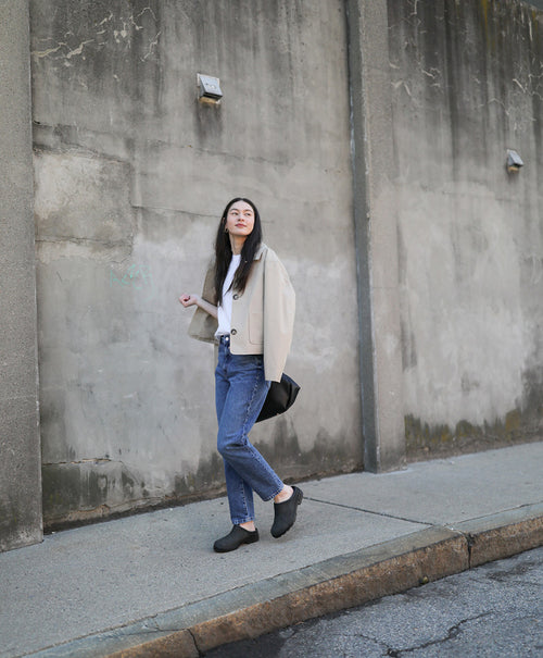 A woman with long dark hair stands on a sidewalk by a weathered concrete wall. She wears a beige jacket, white top, blue jeans, black shoes, and carries a black bag over her shoulder.