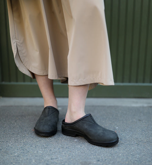 A person stands on a sidewalk wearing a beige skirt or wide-legged pants and black slip-on clogs. The background features a green wall with vertical lines.