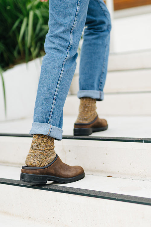 Person wearing blue jeans, brown shoes, and thick speckled socks walking up white stairs with black edges. Green plants are visible in the background.