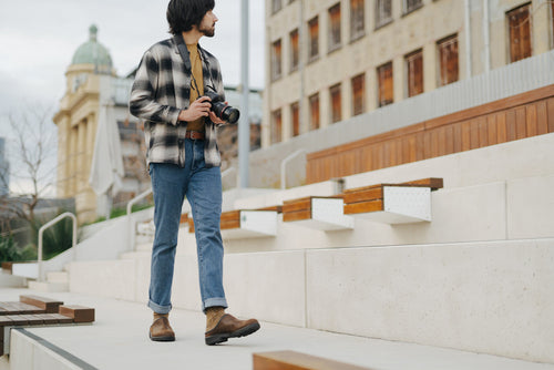 A person wearing jeans, boots, and a plaid shirt walks outdoors on concrete steps holding a camera, with a historic building and trees in the background.