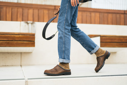 A person wearing blue jeans, brown socks, and brown clogs walks up white steps, holding a pair of black headphones in one hand. Wooden benches and a wooden railing appear in the background.