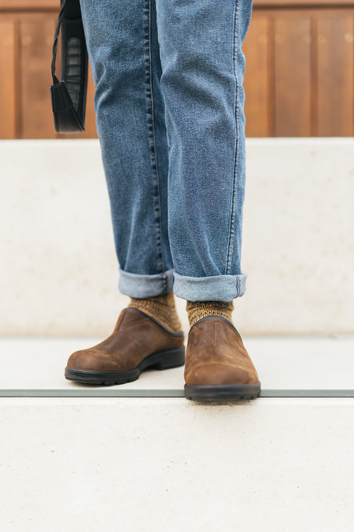 A person wearing cuffed blue jeans, brown slip-on shoes, and patterned socks stands on light-colored steps, holding a black strap in one hand. The background features a wooden wall.