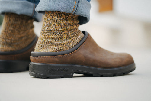 Close-up of a person wearing thick, knit brown socks and brown slip-on clogs with black soles. The person’s jeans are rolled up slightly above the ankles.