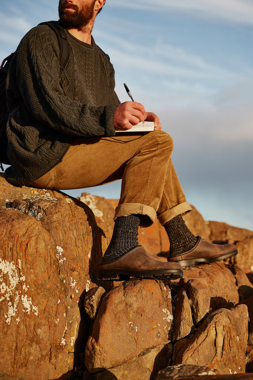 A person with a beard, wearing a dark knit sweater, brown pants, and loafers, sits on large rocks outdoors, writing in a notebook with a pen. The scene is bathed in warm sunlight.