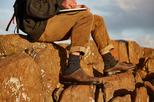 A person in brown pants and dark shoes sits on rocky terrain, writing in a notebook. Only their lower body is visible, with rolled-up pants and textured socks. Warm sunlight highlights the rocks and clothing.