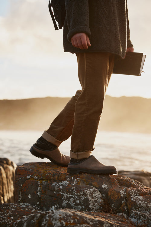A person wearing brown pants, a dark sweater, and boots stands on a mossy rock by the water at sunset, holding a closed notebook in one hand. Only their lower half is visible.