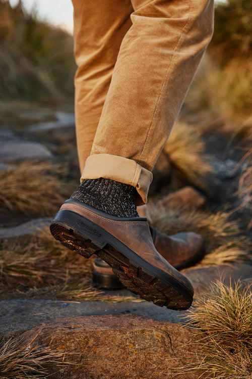 A close-up of a person wearing tan pants, dark wool socks, and brown slip-on shoes walking on a rocky outdoor path, surrounded by grass and plants.