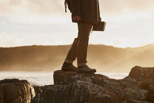 A person stands on rocky terrain near the ocean at sunset, holding a closed book in one hand. Only their lower half is visible, dressed in casual pants and boots, with hills and a soft sky in the background.