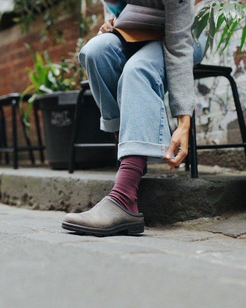 A person sitting on a curb rolls up the cuff of their light blue jeans, wearing maroon socks and brown slip-on shoes. Plants and a brick wall are visible in the background.