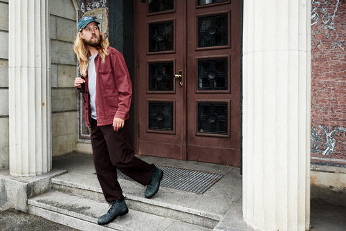 A man with long blond hair and a beard, wearing a green cap, maroon jacket, dark pants, and black boots, walks down stone steps outside a building with large columns and a wooden door.